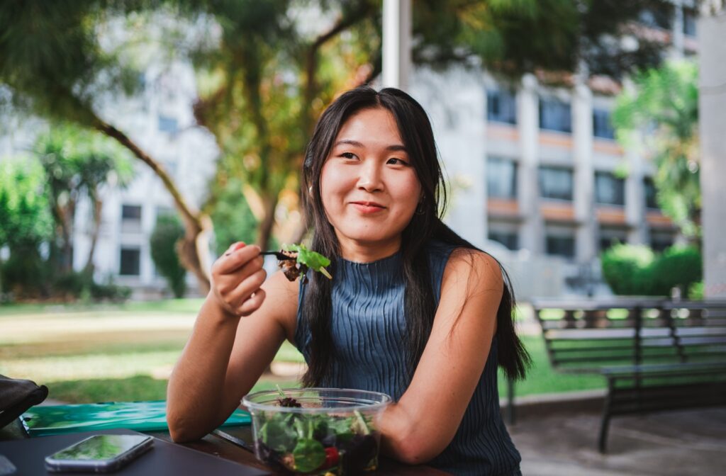 A college-aged woman sits in the park, eating a salad.