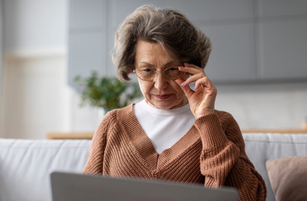 Older woman in brown sweater looking at laptop while holding on to the glasses she is wearing.