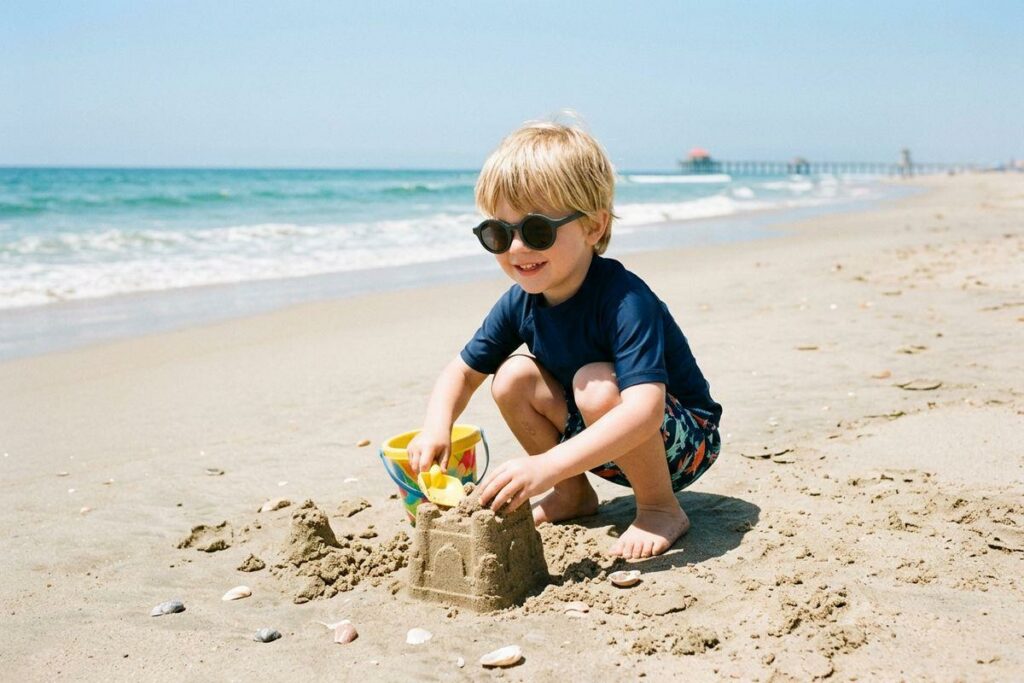 A smiling child in a blue swim shirt and sunglasses squats on a beach, building a sandcastle with a yellow shovel and bucket near the ocean.